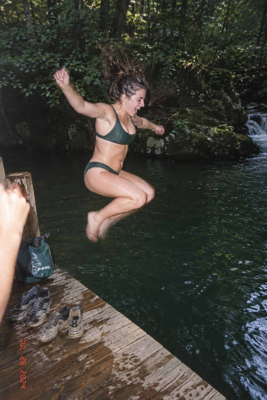 A single candid summer travel photograph with a private-album feel. ONE photo, one frame, one scene, one moment — no collage, no grid. An adult woman in her late twenties caught mid-jump off a weathered wooden dock into a forest creek swimming hole — a transitional, unfinished pose: knees tucked, arms flung out, mouth open in a shout-laugh, hair flying, not looking at the camera. Ordinary vacation swimwear: a dark green sporty two-piece. Realistic skin with pores and goosebumps, wet from a previous jump. Dappled green light through the trees, dark green water, a friend's forearm and phone-free hand at the very edge of the frame as point-of-view evidence. Lived-in details: two pairs of wet sneakers and a dry bag on the dock, wet planks. Shot on a cheap disposable film camera with direct on-camera flash in the shade: limited dynamic range, harsh flash on the subject with a darker background, slight motion blur on the feet, grain, a date stamp in the corner, off-center framing. Only photographic imperfections, never generative ones: correct anatomy, five fingers, no extra limbs.